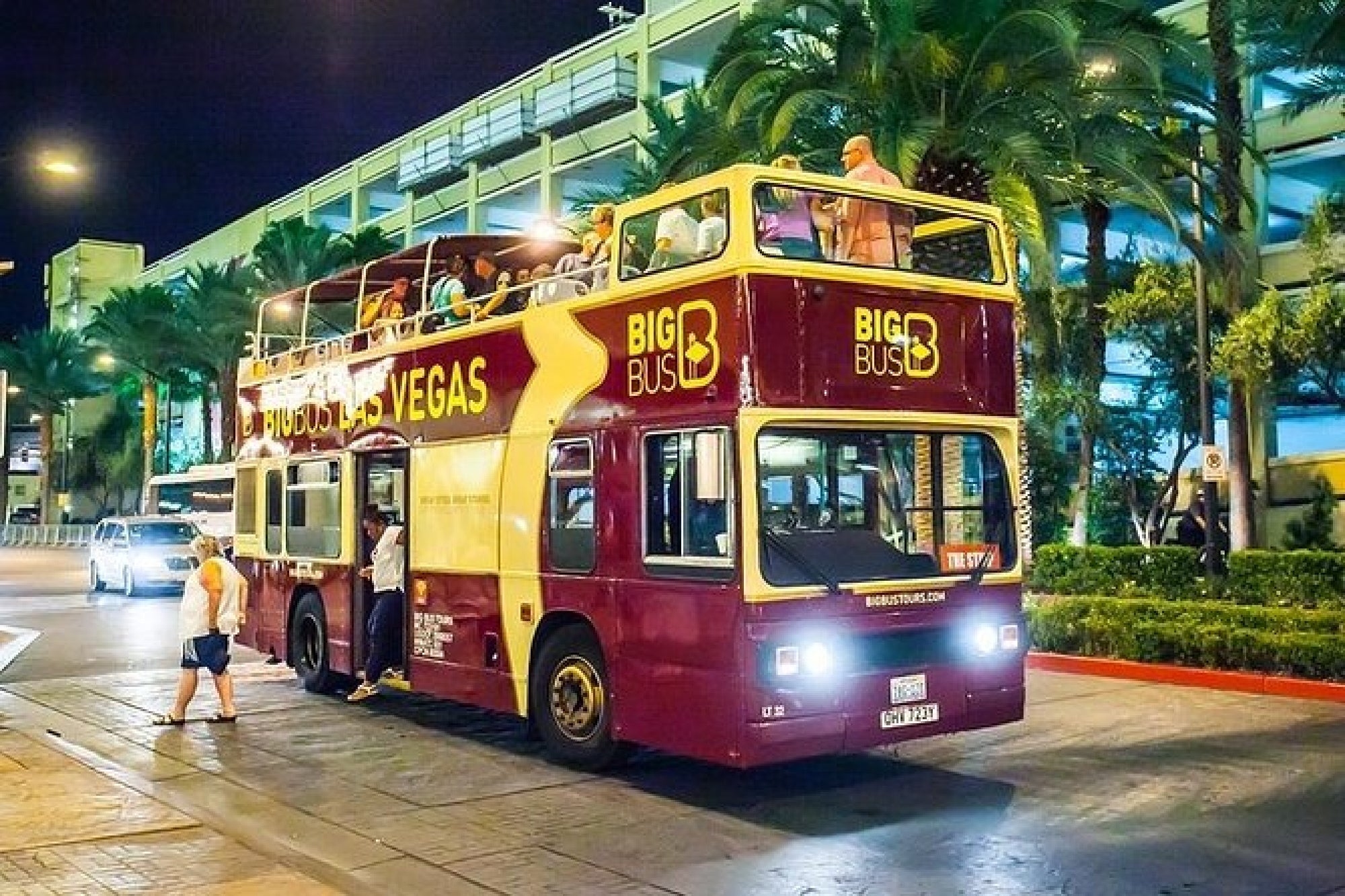 Double-decker bus labeled "Big Bus Las Vegas" with passengers on the top deck, parked on a street at night with palm trees and buildings in the background.