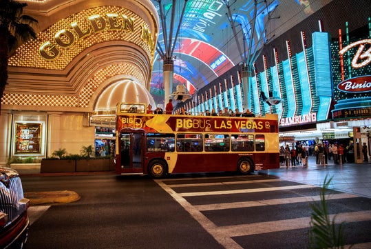 Red double-decker bus labeled "BigBus Las Vegas" on a street with illuminated signs and a casino entrance at night.