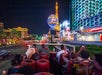 A view of the Las Vegas Strip at night from a double-decker bus. People are taking photos of the bright lights and the Eiffel Tower replica in the background.