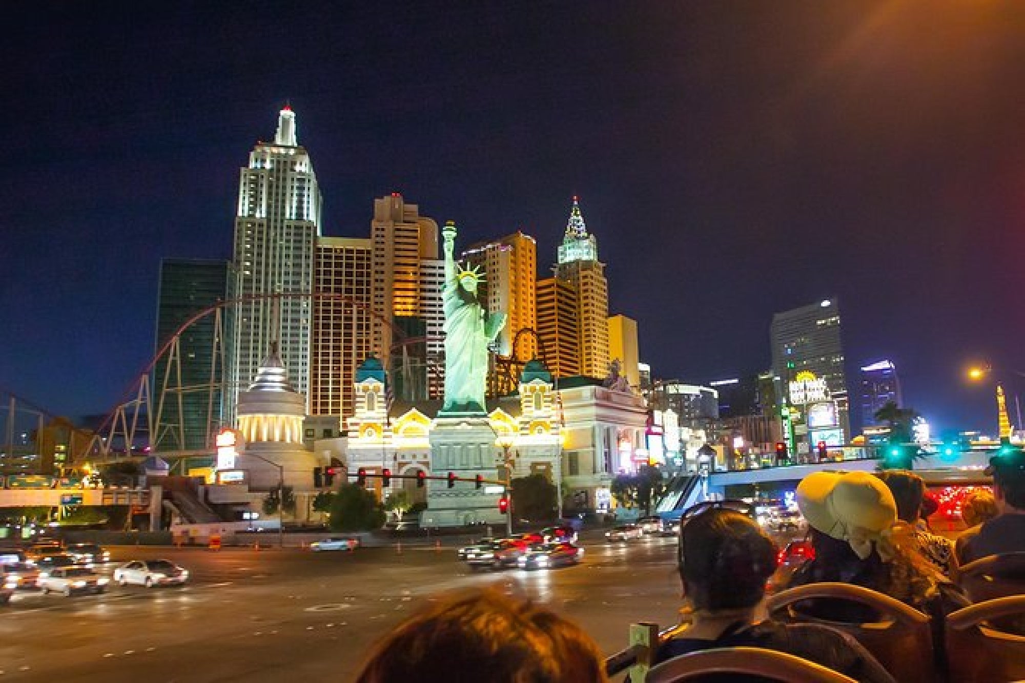A nighttime cityscape featuring a replica of the Statue of Liberty, illuminated buildings, and light traffic on a wide road, with two people in the foreground.