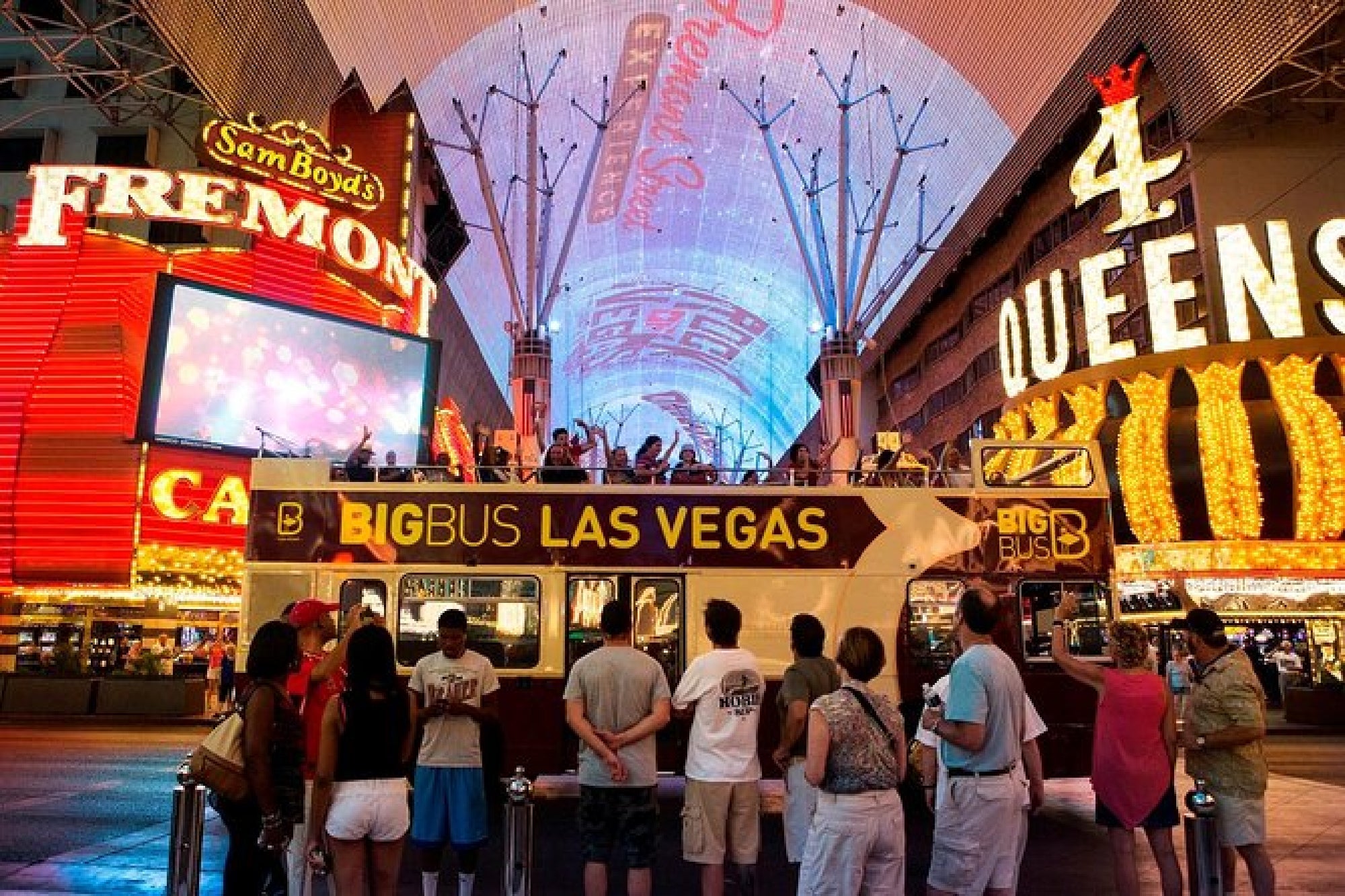 A group of people stands in front of a Big Bus Las Vegas tour bus at night, surrounded by bright neon signs for Fremont and 4 Queens.