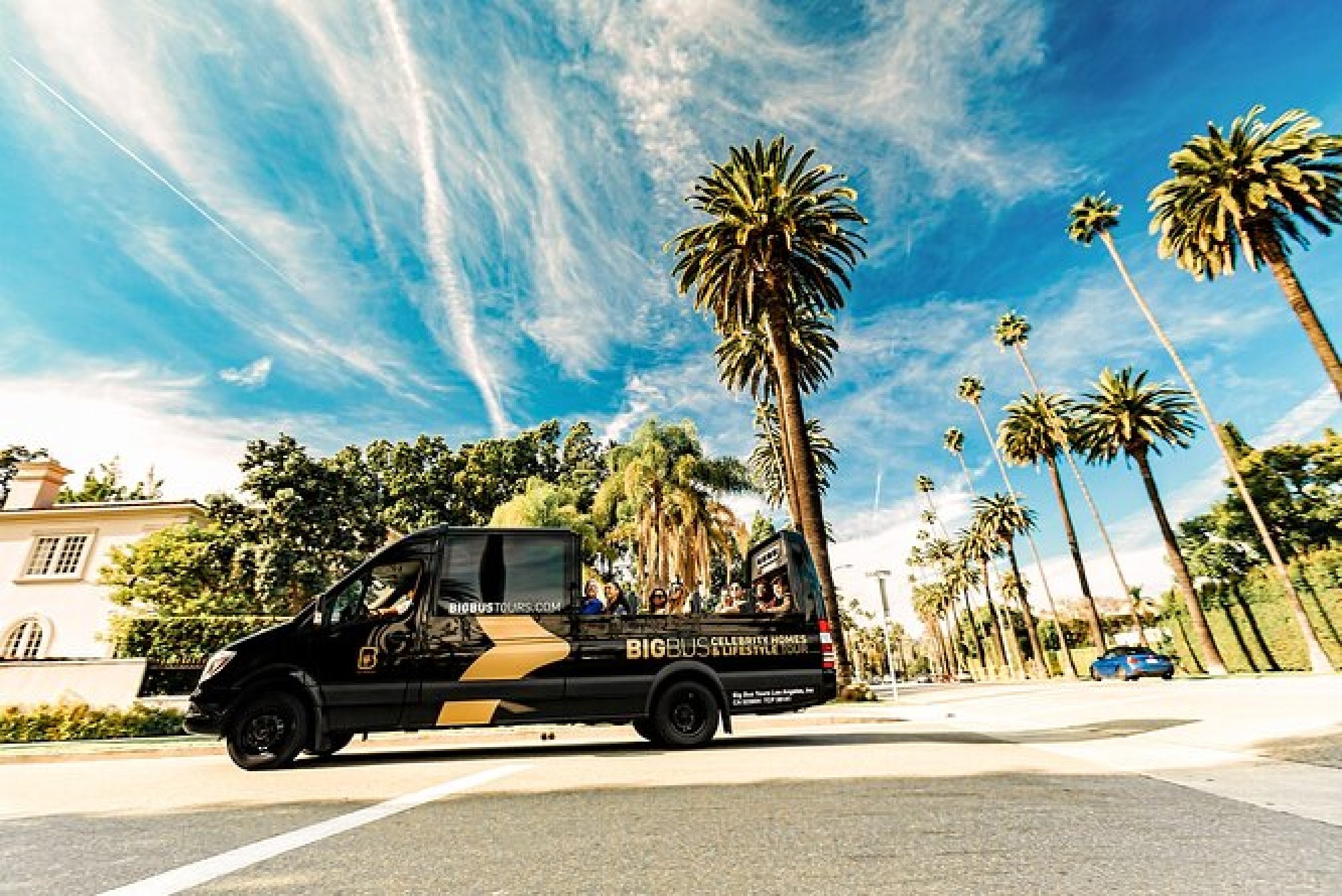 A black tour van drives down a palm tree-lined street under a bright blue sky with wispy clouds.