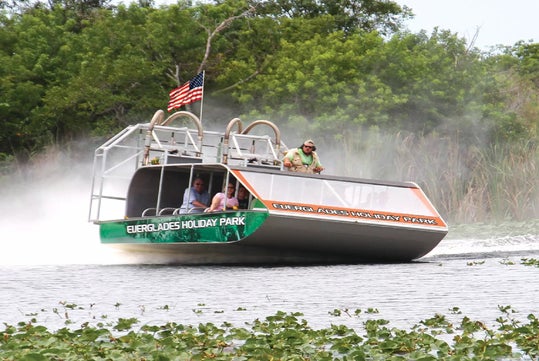 An airboat with people onboard glides through water surrounded by reeds and greenery, displaying an American flag and the text “Everglades Holiday Park” on its side.