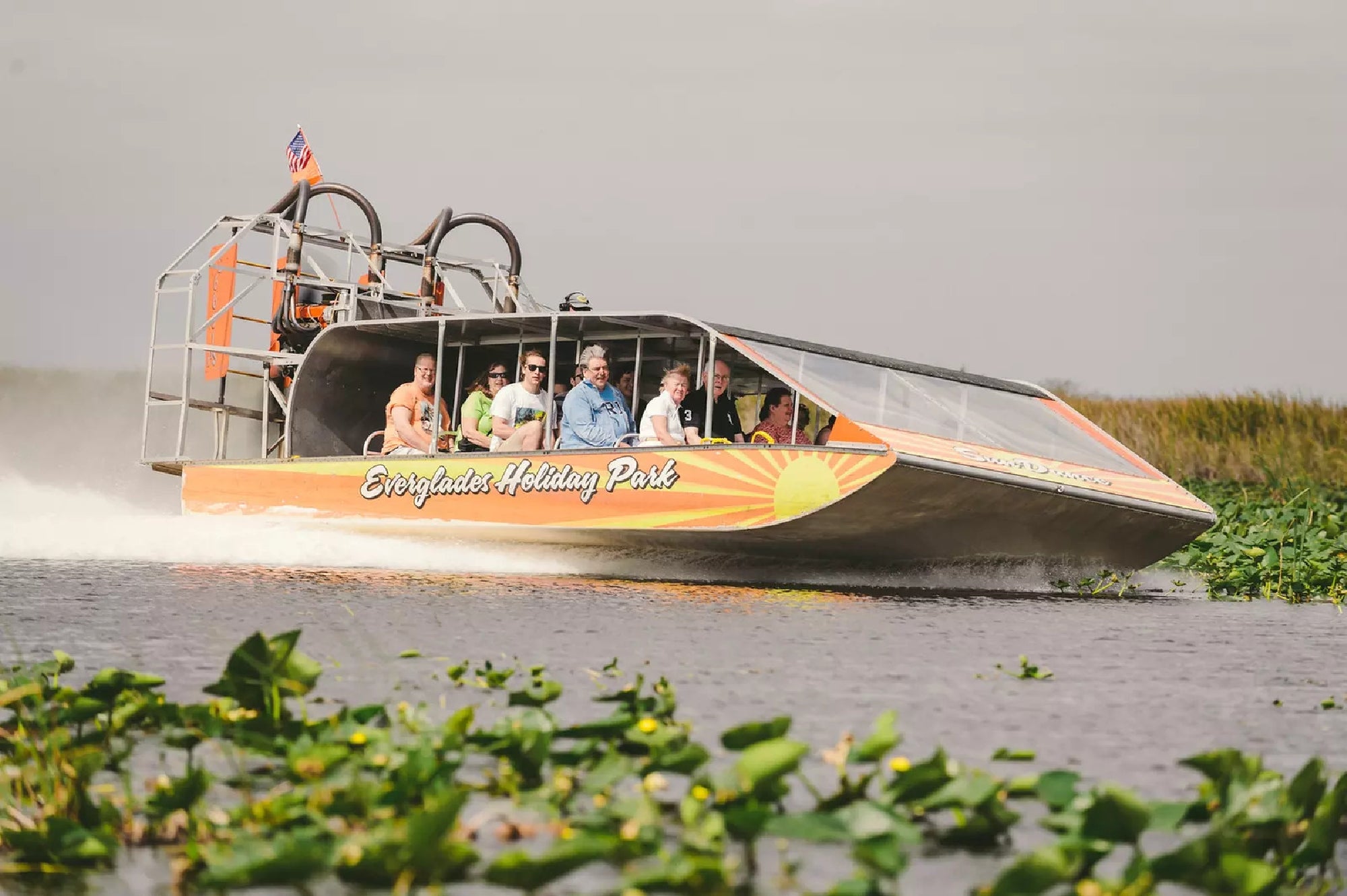 An airboat with passengers glides through wetlands at Everglades Holiday Park, surrounded by water and vegetation on a cloudy day.