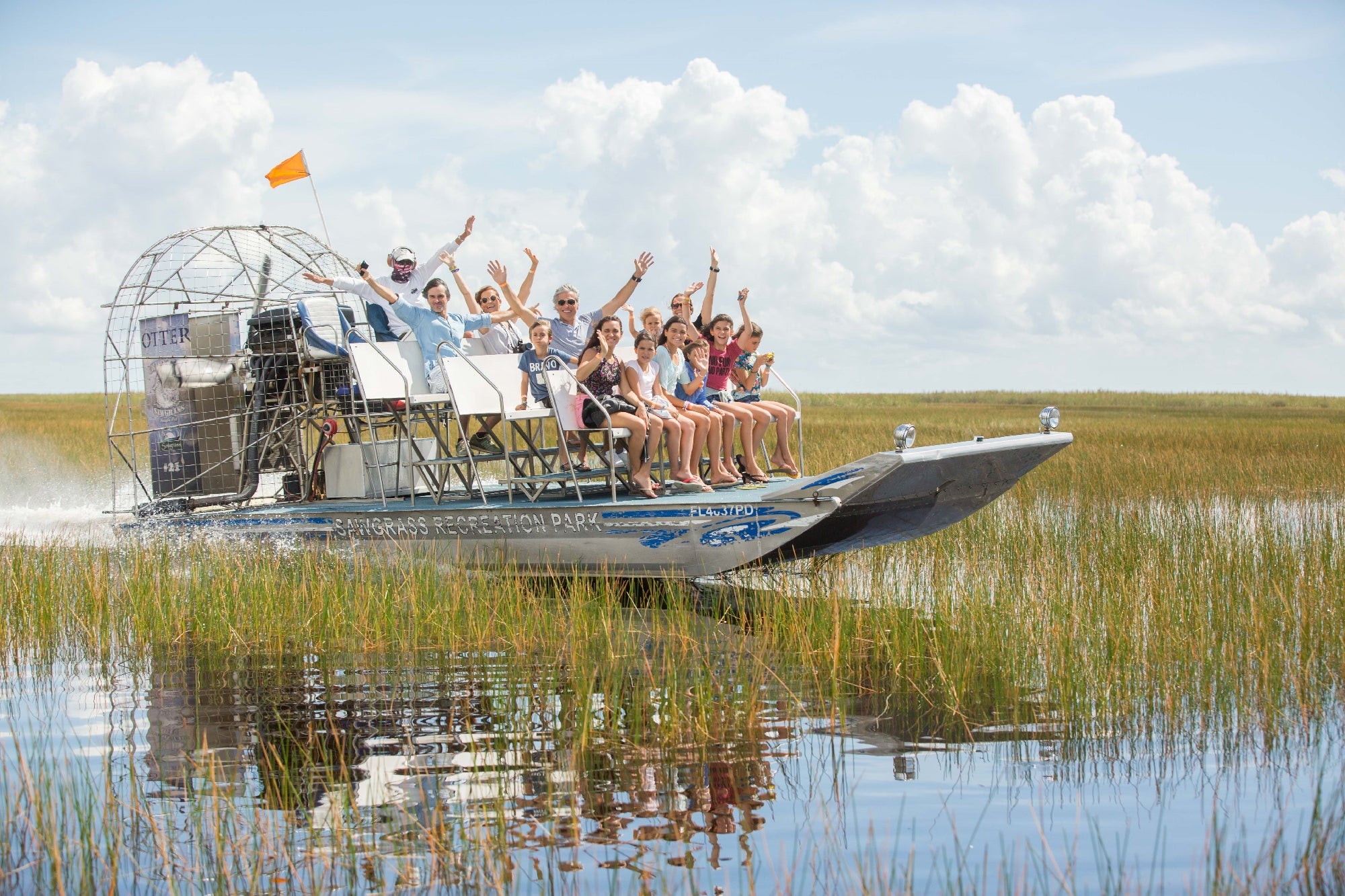 A group of people ride an airboat through grassy wetlands on a sunny day, waving and smiling at the camera.