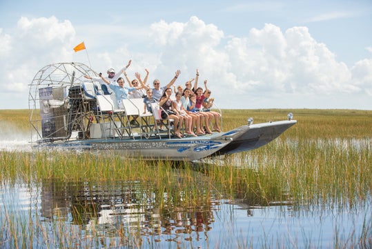 A group of people ride an airboat through grassy wetlands on a sunny day, waving and smiling at the camera.