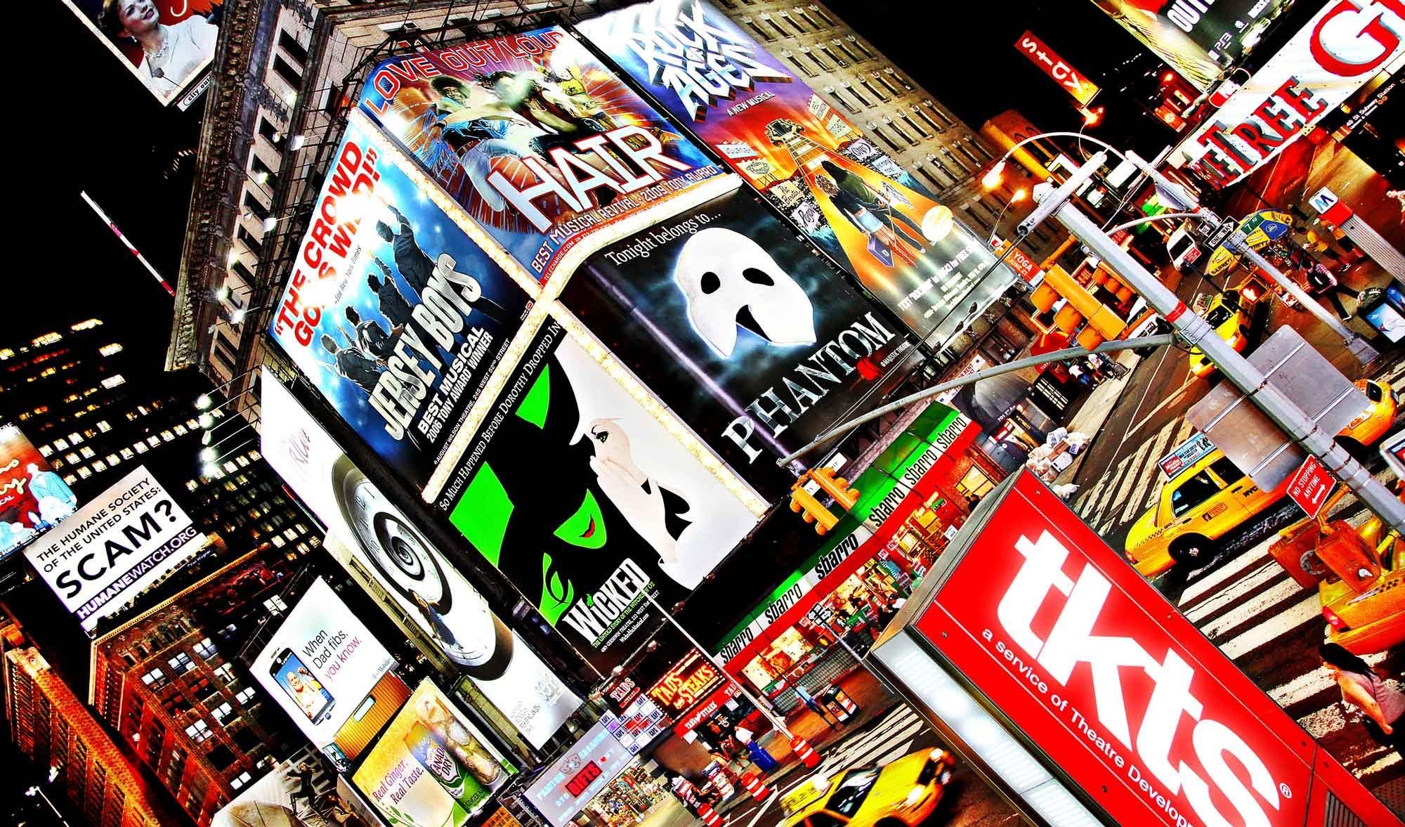 Times Square at night with bright billboards advertising shows like Wicked, Phantom, and Hair, crowded streets, yellow taxis, and people walking.