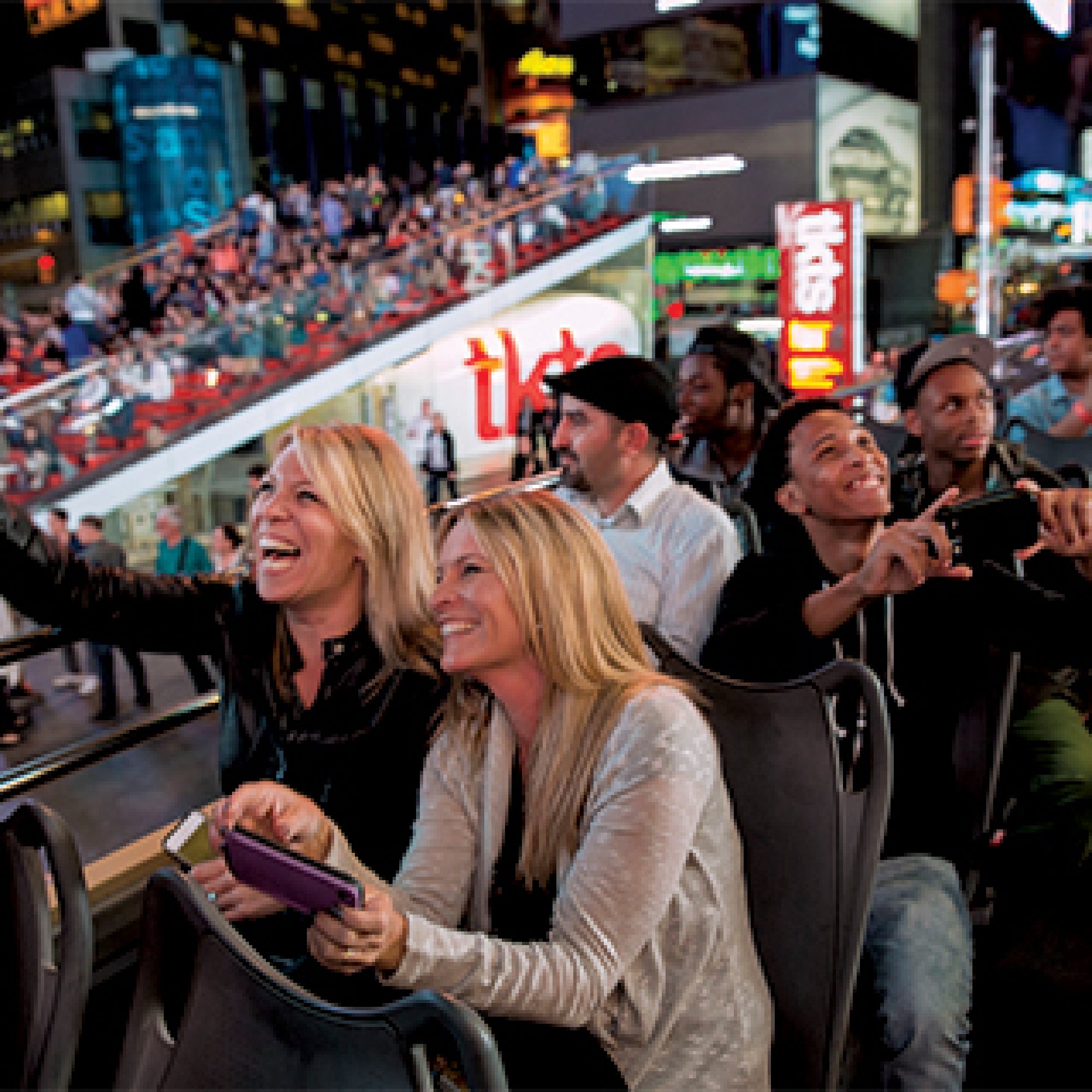 A group of people are seated on an open-top bus at night, taking photos and selfies, with a crowd in the background.