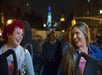 Two women smile and laugh while sitting on a nighttime bus tour with a cityscape in the background. Other passengers can be seen seated behind them.