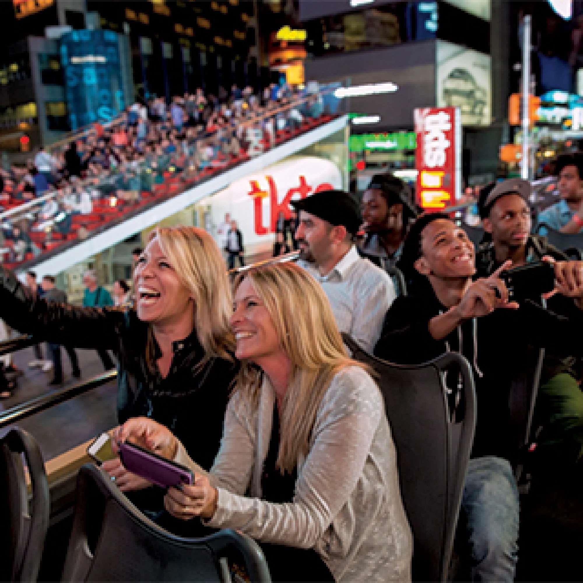 People sit on the upper deck of a tour bus at night, smiling and taking photos, with a crowd and bright city lights in the background.