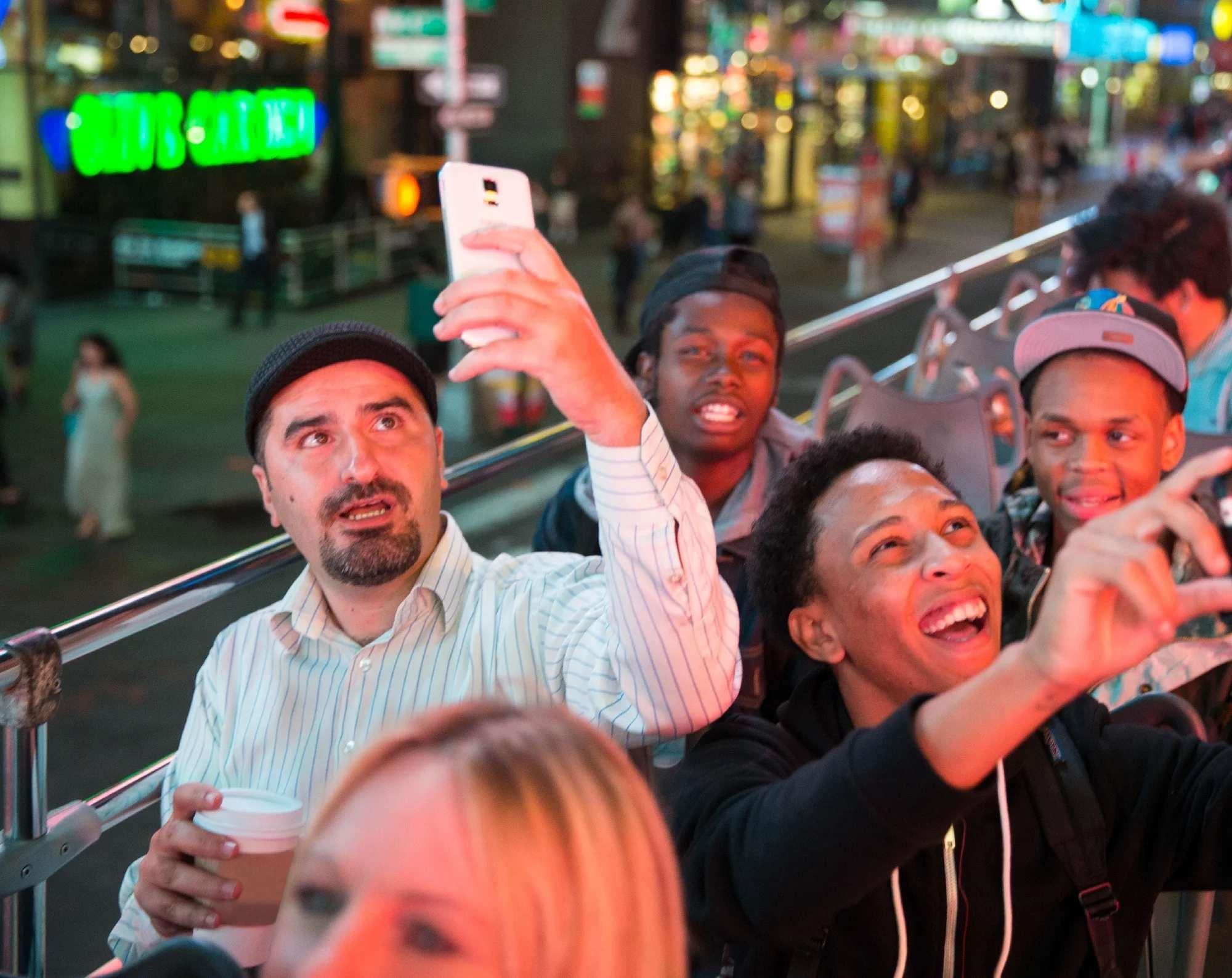 A group of people on an open-top bus at night, with one man holding a phone for a selfie and others smiling and pointing, city lights visible in the background.