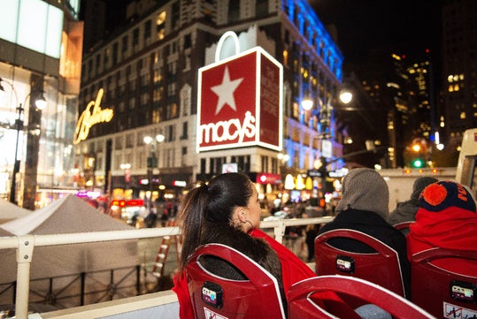 People on an open-top bus passing by Macy's in a city at night, with illuminated signs and buildings in the background.