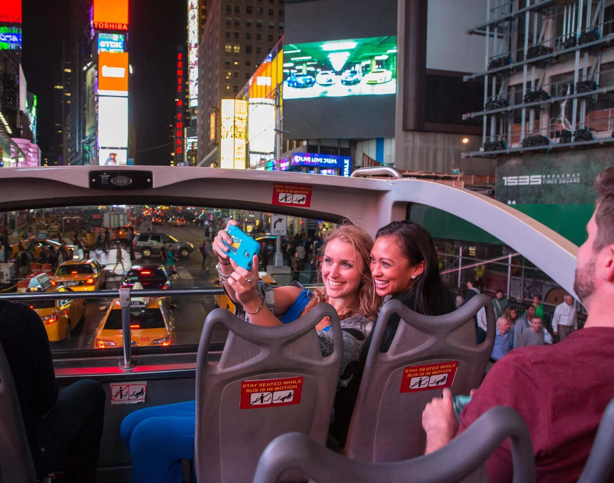 Two women take a selfie on the upper deck of a tour bus at night in Times Square, New York City, with bright billboards and traffic in the background.