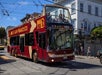 A red double-decker tour bus labeled "Big Bus San Francisco" drives through a city street with passengers seated on the open upper deck.