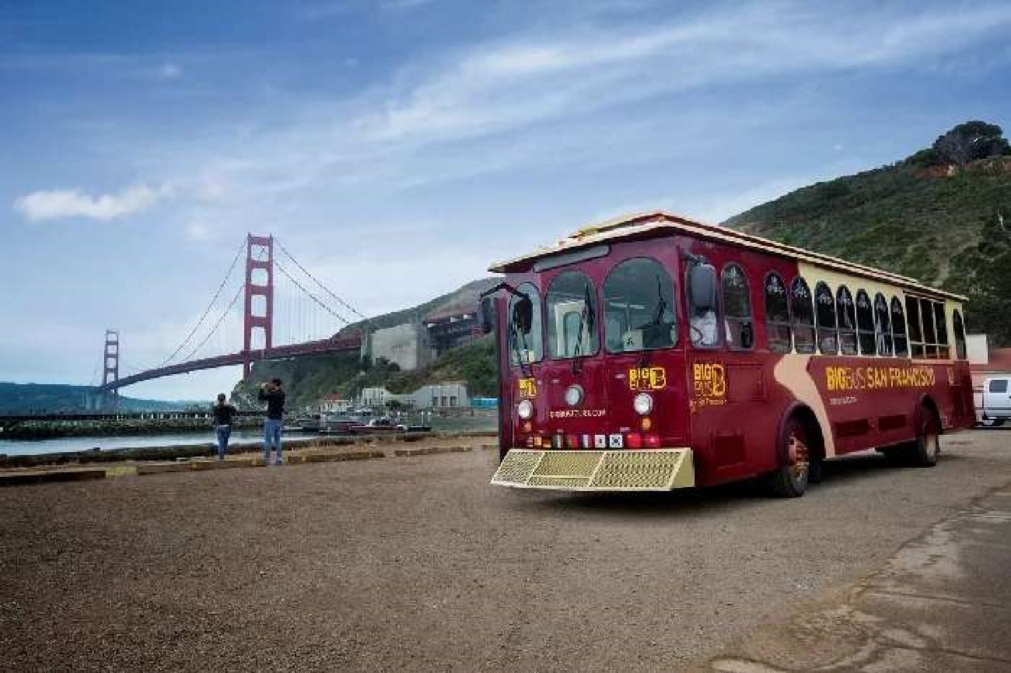 A red and yellow San Francisco tour trolley is parked near the waterfront, with the Golden Gate Bridge and two people visible in the background.