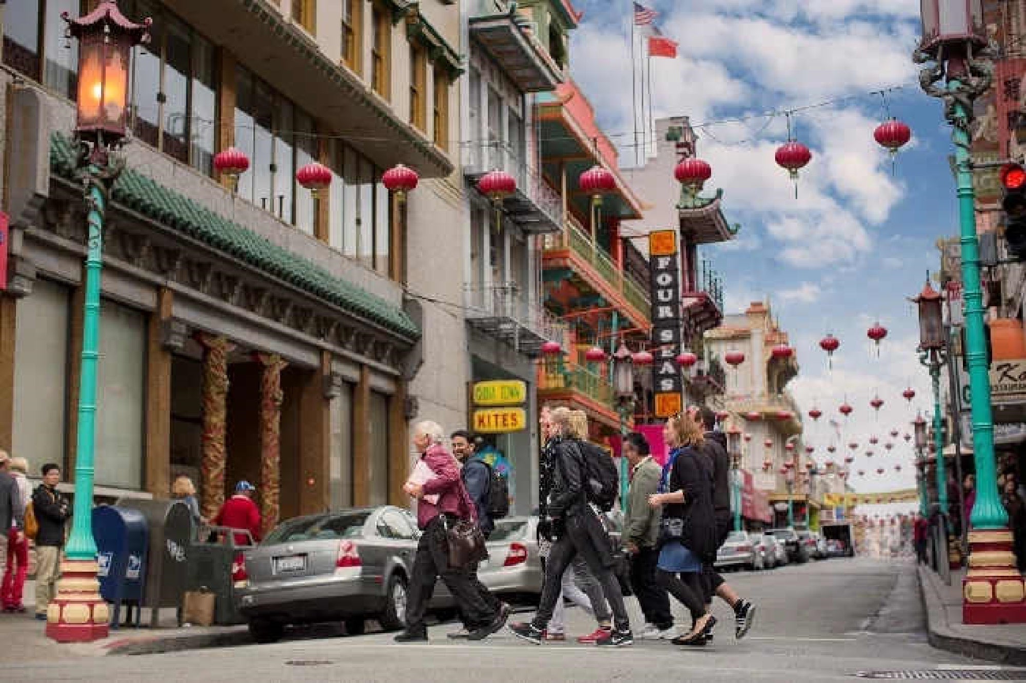 A group of people cross a street in a city’s Chinatown, surrounded by buildings with lanterns, signs, and ornate architectural details.