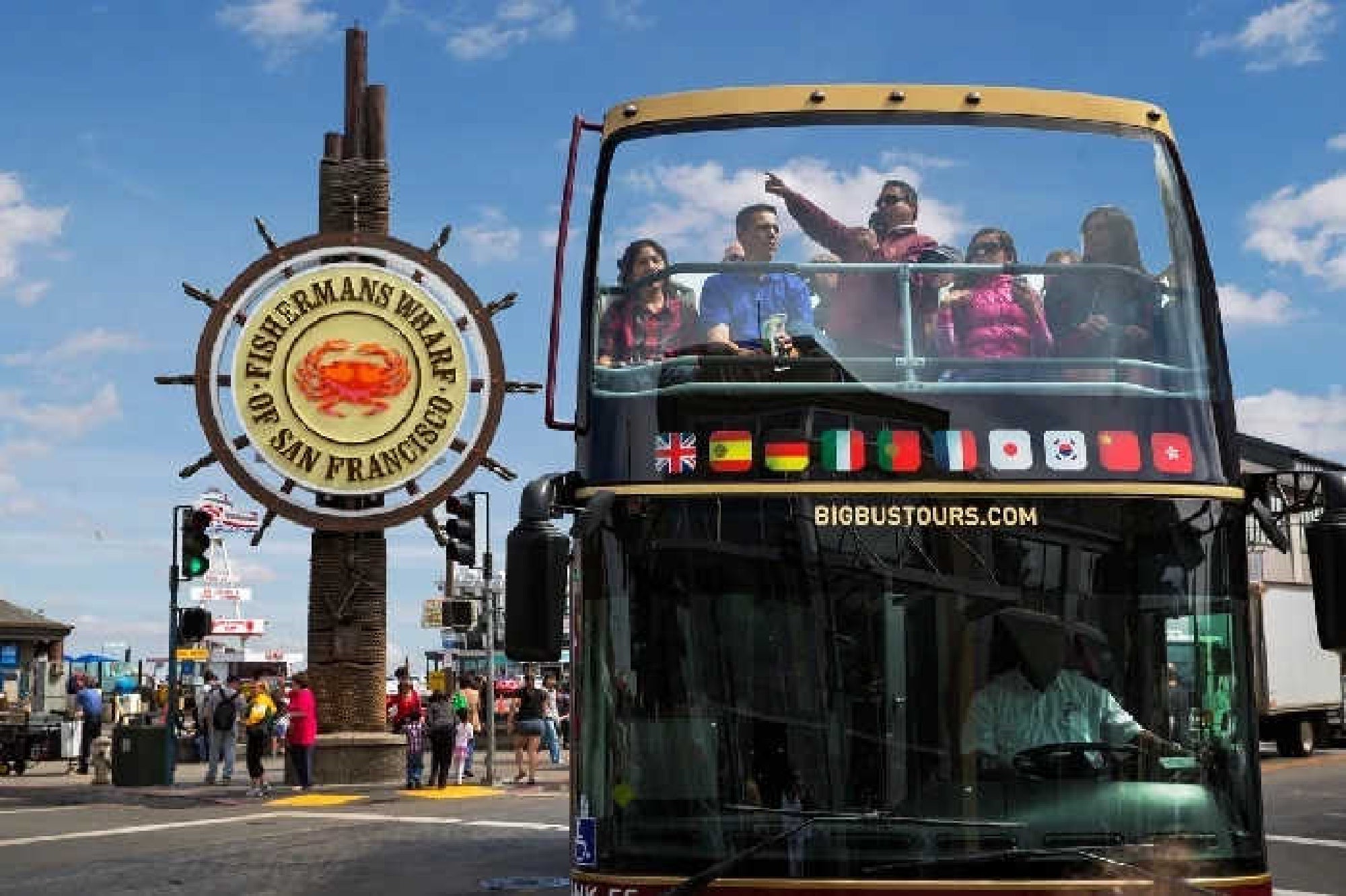 A group of people ride on the upper deck of a sightseeing bus near the Fisherman's Wharf sign in San Francisco on a sunny day.