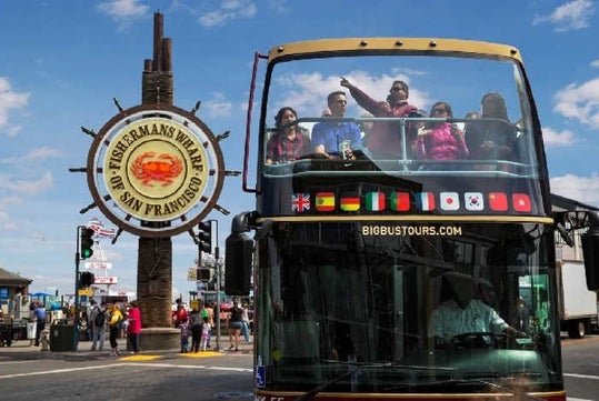 A group of people ride on the upper deck of a sightseeing bus near the Fisherman's Wharf sign in San Francisco on a sunny day.