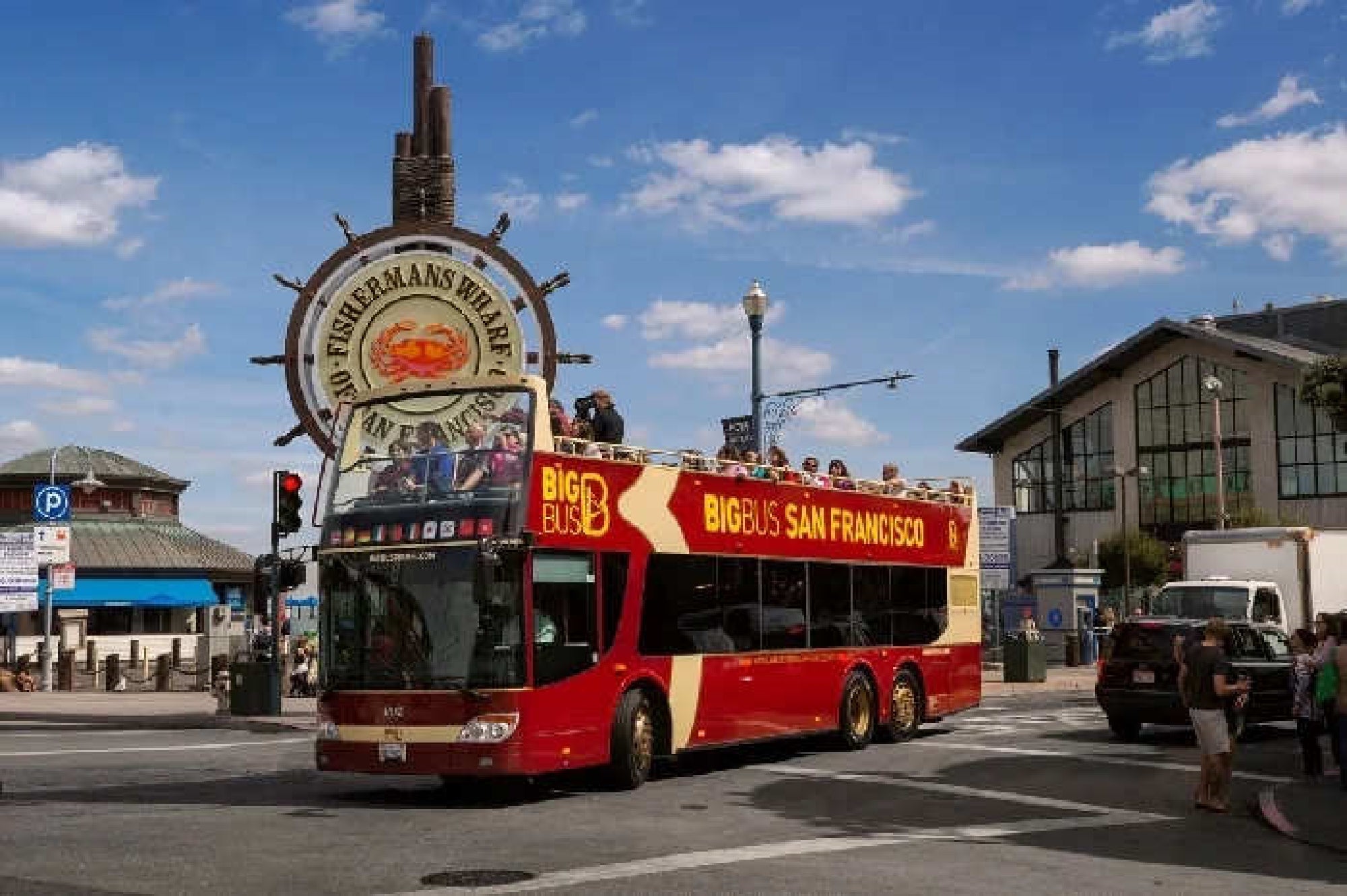A red double-decker tour bus labeled “Big Bus San Francisco” drives past the Fisherman's Wharf sign on a sunny day.