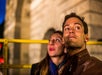 A man and a woman wearing earbuds look up while standing outdoors at night, with a blurred stone building in the background.