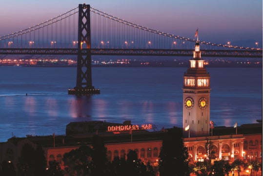 The San Francisco Ferry Building and clock tower are illuminated at dusk with the Bay Bridge and blue water in the background.