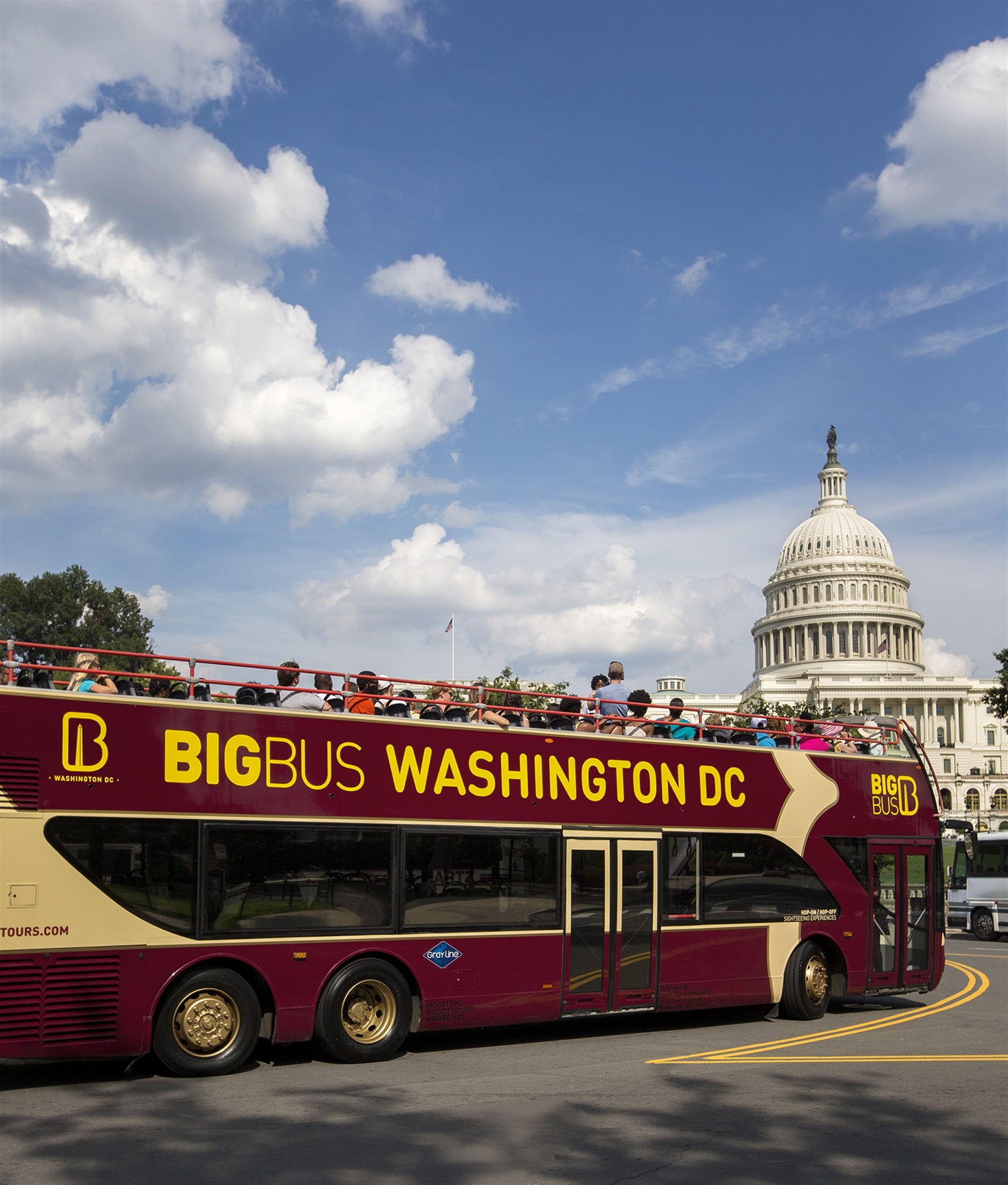 A double-decker tour bus labeled "Big Bus Washington DC" drives past the United States Capitol under a blue sky with scattered clouds.