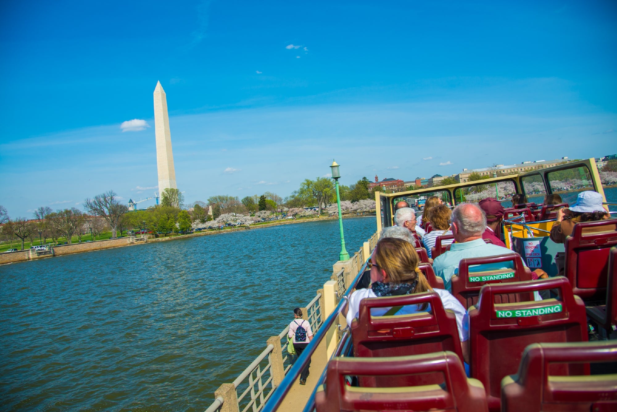 Tourists on an open-top bus ride along a waterfront with the Washington Monument visible under a clear blue sky.