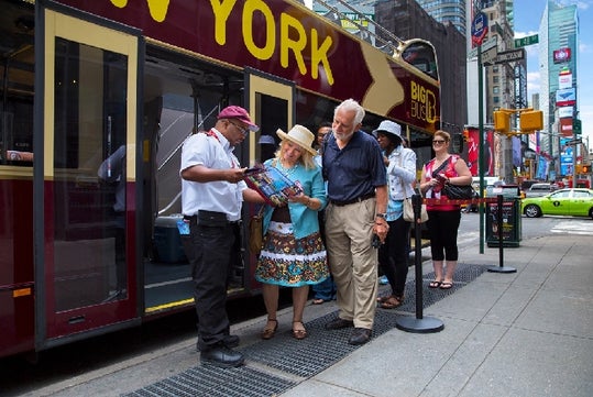 A group of people stand on a sidewalk near a tour bus in New York as a staff member answers questions from an older couple holding a map.