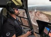 A pilot wearing sunglasses and a headset flies a helicopter over an active volcano, with smoke rising from the crater below.