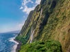 Steep coastal cliffs with cascading waterfalls meet the ocean under a blue sky and scattered clouds.