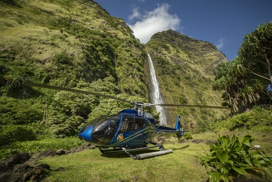 A Helicopter sitting on a clearing in front of a waterfall and cliff on the Big Island Spectacular Helicopter Tour in Hawaii USA.
