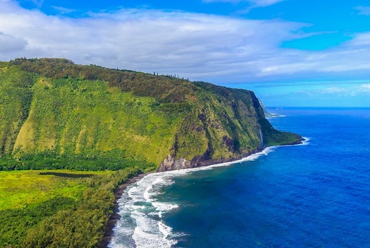 Aerial view of a green cliffside meeting the blue ocean, with waves breaking along the shoreline under a partly cloudy sky.