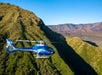 A blue helicopter flies over green, hilly terrain with mountains in the background under a clear blue sky.