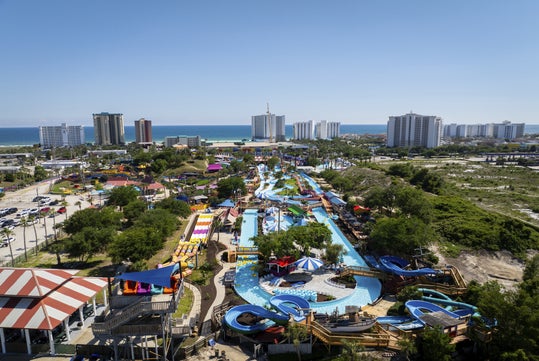 Aerial view of a water park with pools, slides, and colorful structures, surrounded by trees, parking lots, and tall buildings near a beach and ocean.
