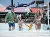 A family of four, all wearing swimwear, walks and laughs together in shallow water at a water park with slides and colorful structures in the background.