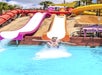 A person splashes into a pool from the end of an orange and white water slide at an outdoor water park on a sunny day.