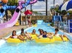 A group of people float on yellow inner tubes in a lazy river at a water park, with water slides and walkways in the background.