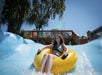 A young woman rides a yellow inflatable tube down a blue water slide at a water park on a sunny day.