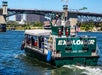 A tour boat labeled "Explorer" navigates a river with a bridge and cityscape in the background. A few passengers are seated under a canopy.