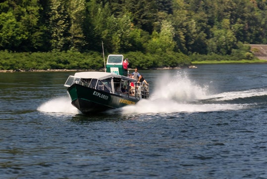 A speedboat named "Explorer" moves rapidly on a river, creating a large wake. Trees line the riverbank in the background.