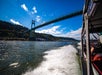 View from a boat speeding under a suspension bridge over a river, with green hilly landscape in the background and a blue sky above.