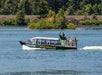 A boat named "Explorer" with "BigfootCruise.com" on the side travels on a river. Passengers are seated under a canopy, while some stand at the front. Trees and grass are in the background.