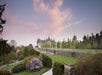 A large historic mansion sits in the distance amid landscaped gardens and green lawns under a pink and blue sky at sunset.