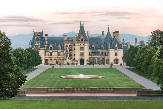 A large, ornate mansion with multiple chimneys and turrets stands behind a circular fountain and a manicured lawn, framed by trees and mountains in the background.