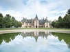 Large historic mansion with turrets and tall windows, reflected in a round pond, surrounded by green trees under a partly cloudy sky.