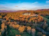 Aerial view of a large mansion surrounded by colorful autumn trees and rolling hills under a blue sky with scattered clouds.