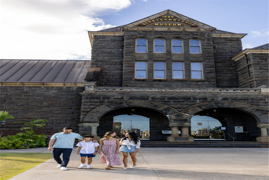 A family of four walks in front of a historic stone museum building with arched entryways and the sign "1899 Bernice Pauahi Bishop Museum" above the entrance.