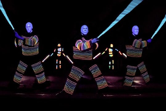 Performers in colorful, glowing costumes and masks hold illuminated staffs in a dark setting.