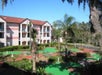 Three-story apartment building with balconies surrounded by manicured greenery and a mini-golf course under a clear blue sky.