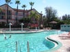 Outdoor pool with swimmers, surrounded by palm trees. Hotel building in the background with balconies. Lounge chairs and umbrellas are set up around the pool's edge.